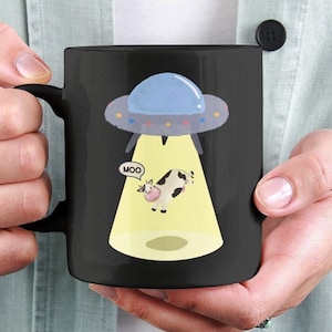 Op de afbeelding: Zwarte koffiemok met een cartoon-koe die wordt ontvoerd door een UFO. De koe zegt "boe" in een spraakballon.