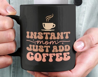 Puede incluir: Una taza de café negra con un icono de taza de café blanco y el texto "Instant mom - just add coffee" en rosa.