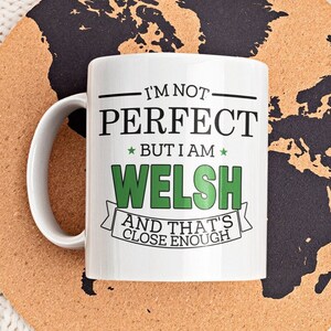May include: White ceramic mug with a black and green design that reads "I'm not perfect but I am Welsh and that's close enough". The mug is sitting on a cork coaster with a black world map design.