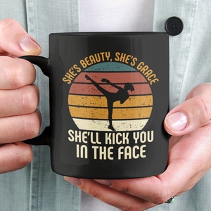 May include: Black coffee mug with a retro-style graphic of a woman doing a karate kick. The text reads "She's beauty, she's grace, she'll kick you in the face."