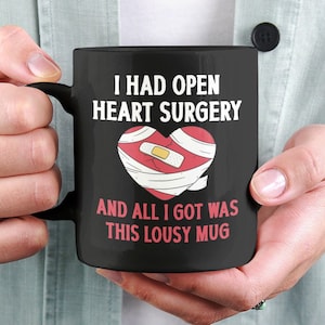 Puede incluir: Taza de café negra con texto blanco que dice "Tuve una cirugía a corazón abierto y todo lo que obtuve fue esta taza de mala muerte". Un corazón rojo con un vendaje está ilustrado en la taza.