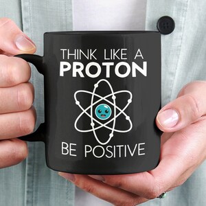 Może przedstawiać: Czarny kubek do kawy z białym tekstem, który brzmi "Myśl jak proton, bądź pozytywny". Kubek przedstawia stylizowany atom z niebieską uśmiechniętą twarzą w środku.