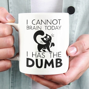 May include: White ceramic mug with the text "I CANNOT BRAIN TODAY I HAS THE DUMB" and a black cat illustration. The mug is held by a person wearing a light green shirt.