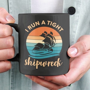 May include: Black coffee mug with a white graphic design that says "I run a tight shipwreck" with a vintage sunset and a silhouette of a ship.