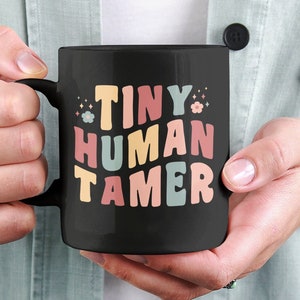 Peut inclure: Une tasse à café noire avec le texte "TINY HUMAN TAMER" en lettres colorées de style rétro. La tasse est tenue par une personne.