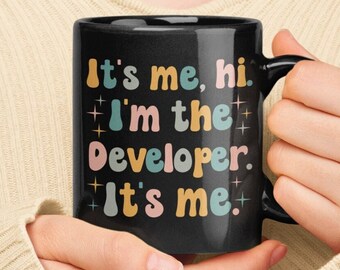 Puede incluir: Una taza de café negra con un diseño blanco que dice "It's me, hi. I'm the developer. It's me." en letras coloridas de estilo retro.