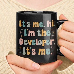 May include: A black coffee mug with a white design that says "It's me, hi. I'm the developer. It's me." in colorful, retro-style lettering.