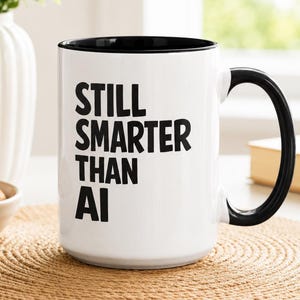 Może przedstawiać: Biały ceramiczny kubek z czarnym wnętrzem i czarnym uchwytem. Na zewnętrznej stronie znajduje się czarny napis "STILL SMARTER THAN AI" wydrukowany pogrubioną czcionką bezszeryfową. Kubek ma cylindryczny korpus i zakrzywiony uchwyt po prawej stronie.