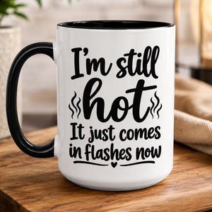Op de afbeelding: Een witte keramische mok met een zwarte binnenkant en handvat. De voorzijde bevat zwarte tekst die luidt: I'm still hot, It just comes in flashes now, met kleine stoompictogrammen boven het woord hot en een klein hartje onder de tekst.