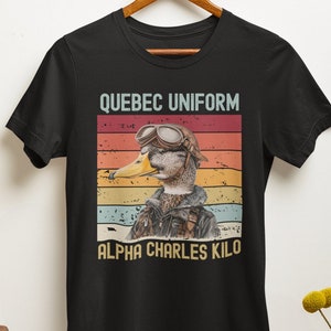 May include: Black t-shirt with a vintage-style graphic of a duck wearing aviator goggles and a flight suit. The text "Quebec Uniform Alpha Charles Kilo" is printed below the graphic.