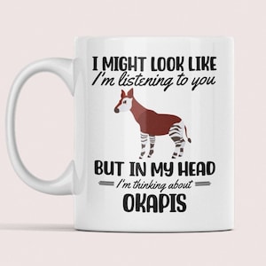 May include: White ceramic mug with a black and brown okapi illustration. The mug has the text "I might look like I'm listening to you, but in my head I'm thinking about okapis."