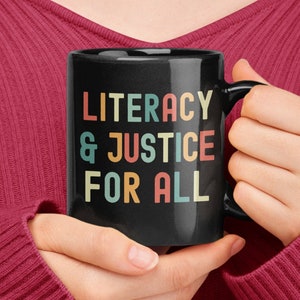 May include: A black coffee mug with a white inscription that reads "Literacy & Justice For All" in colourful retro lettering.