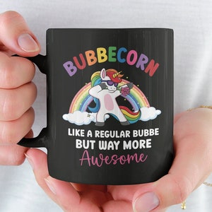 May include: Black coffee mug with a white unicorn wearing sunglasses and a rainbow behind it. The text on the mug reads "Bubblecorn Like a regular bubbe but way more Awesome".