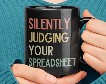 May include: A black coffee mug with the text "SILENTLY JUDGING YOUR SPREADSHEET" in a rainbow colour scheme.