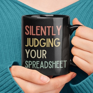 以下が含まれることがあります： 虹色のカラーリングで「SILENTLY JUDGING YOUR SPREADSHEET」と書かれた黒いコーヒーカップです。
