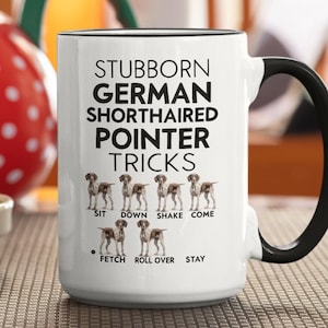 May include: White ceramic coffee mug with a black handle. The mug has a black rim and black text that reads "Stubborn German Shorthaired Pointer Tricks".  The text is surrounded by illustrations of German Shorthaired Pointers performing tricks, including "sit", "down", "shake", "come", "fetch", "roll over", and "stay".