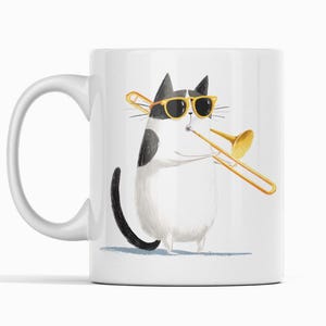 Puede incluir: Taza de cerámica blanca con una ilustración humorística de un gato blanco y negro con gafas de sol tocando un trombón dorado. La taza tiene un asa curva y está sobre un fondo blanco.