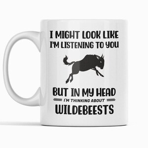 Może przedstawiać: Biały ceramiczny kubek z czarną sylwetką skaczącego gnu. Tekst brzmi: "Mogę wyglądać tak, jakbym słuchał, ale w mojej głowie myślę o gnu."