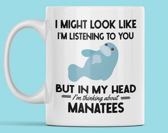 May include: White ceramic coffee mug with a black and white graphic of a manatee. The text on the mug reads "I might look like I'm listening to you, but in my head I'm thinking about manatees."
