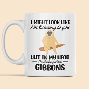 May include: White ceramic mug with a black and white illustration of a gibbon sitting on a branch. The text on the mug reads "I might look like I'm listening to you, but in my head I'm thinking about gibbons."