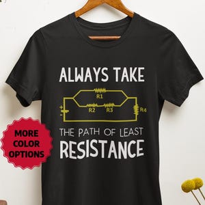 Op de afbeelding: Zwart T-shirt met de tekst "ALWAYS TAKE THE PATH OF LEAST RESISTANCE" en een geel circuitdiagram. Een rode sticker met "MORE COLOR OPTIONS" staat links. Het shirt hangt aan een houten hanger.