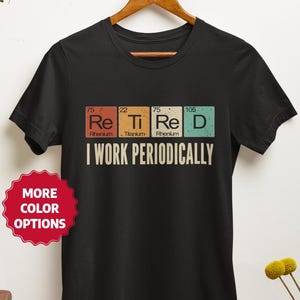 May include: Black t-shirt with the text "I WORK PERIODICALLY" and the periodic table elements Re, Ti, Re, and D. The shirt is hanging on a wooden hanger. A red sticker with "MORE COLOR OPTIONS" is on the left.