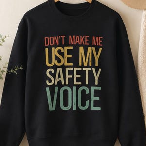 以下が含まれることがあります： 「DON'T MAKE ME USE MY SAFETY VOICE」というテキストが、赤、黄、ライトグリーンのスタックされたマルチカラーデザインでプリントされた黒の長袖スウェットシャツ。文字にはヴィンテージ風の使い古された質感が施されています。