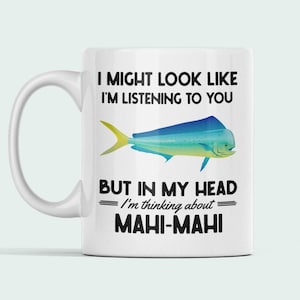 以下が含まれることがあります： 白磁のコーヒーカップに、黒と白の mahi-mahi の魚のグラフィックが描かれています。カップには、「私は聞いているように見えるかもしれませんが、私の頭の中では mahi-mahi のことを考えています。」というテキストが書かれています。