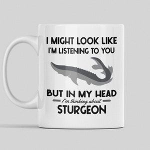 May include: White ceramic coffee mug with a black and gray illustration of a sturgeon fish. The text on the mug reads "I might look like I'm listening to you, but in my head I'm thinking about sturgeon."