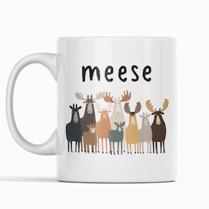 Könnte beinhalten: Weiße Keramiktasse mit dem Wort "meese" in schwarzer Schrift. Darunter befinden sich Cartoon-Elche in verschiedenen Farben, darunter Schwarz, Braun und Beige. Die Tasse hat einen weißen Henkel und steht vor einem weißen Hintergrund.