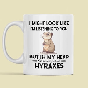 Puede incluir: Taza de café de cerámica blanca con una ilustración en blanco y negro de un animal pequeño. El texto en la taza dice "I might look like I'm listening to you but in my head I'm thinking about HYRAXES".