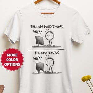 May include: White t-shirt with a humorous black and white graphic. The graphic depicts a stick figure at a computer with the text "THE CODE DOESN'T WORK WHY?" and "THE CODE WORKS WHY?" A red sticker with "MORE COLOR OPTIONS" is visible.