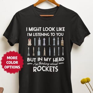 Op de afbeelding: Zwart t-shirt met de tekst "I MIGHT LOOK LIKE I'M LISTENING TO YOU BUT IN MY HEAD I'm thinking about ROCKETS." Het shirt heeft een afbeelding van verschillende raketten. Een rode sticker zegt "MORE COLOR OPTIONS."