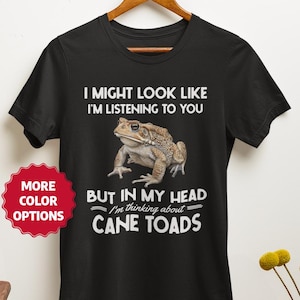 Puede incluir: Camiseta negra con un gráfico de un sapo y el texto: "I MIGHT LOOK LIKE I'M LISTENING TO YOU BUT IN MY HEAD I'm thinking about CANE TOADS." Una pegatina roja con "MORE COLOR OPTIONS" está a la izquierda.