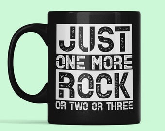 May include: Black ceramic coffee mug with white text that reads "JUST ONE MORE ROCK OR TWO OR THREE".