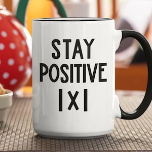 Puede incluir: Taza de cerámica blanca con borde y asa negros. La taza tiene texto negro que dice "STAY POSITIVE IXI".