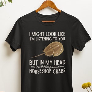 May include: Black t-shirt with the text "I MIGHT LOOK LIKE I'M LISTENING TO YOU BUT IN MY HEAD I'm thinking about HORSESHOE CRABS." The shirt features a horseshoe crab graphic. The shirt is hanging on a wooden hanger.