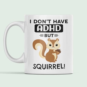 May include: White ceramic mug with a black and white design. The text reads "I don't have ADHD but= SQUIRREL!" and features a cartoon squirrel holding an acorn.