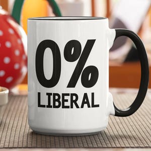 May include: White ceramic mug with a black handle and rim. The mug features the text "0% LIBERAL" in bold black letters. A red polka-dot teapot and a bowl of cookies are in the background.