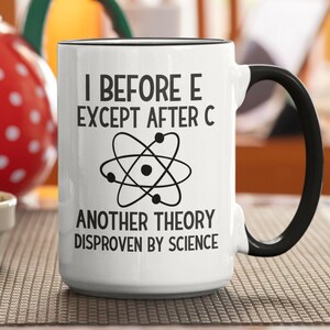 May include: White ceramic mug with a black handle and rim. The mug features the text "I BEFORE E EXCEPT AFTER C" and "ANOTHER THEORY DISPROVEN BY SCIENCE" with an atom symbol. The mug is on a brown surface.