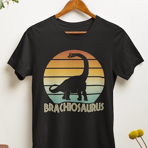 May include: Black t-shirt featuring a vintage-style graphic of a Brachiosaurus dinosaur silhouette against a sunset backdrop. The text "Brachiosaurus" is printed beneath the dinosaur.