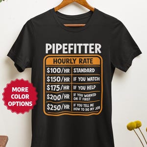 May include: Black t-shirt with the text "PIPEFITTER HOURLY RATE" and a list of hourly rates based on the level of assistance. The shirt is hanging on a wooden hanger.