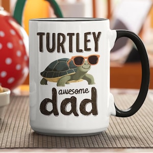 May include: White ceramic mug with a black handle and rim, featuring the text "TURTLEY awesome dad" and a cartoon turtle wearing sunglasses. The mug is on a patterned surface.
