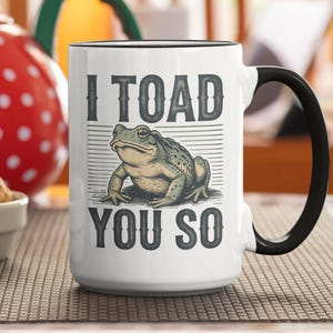 May include: White ceramic mug with a black handle and rim. The mug features the text "I TOAD YOU SO" with a detailed illustration of a frog. The mug is on a brown surface.
