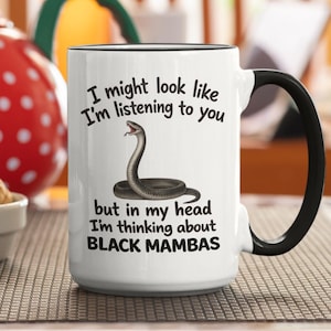 Op de afbeelding: Witte keramische mok met een zwart handvat en rand. De mok heeft de tekst "I might look like I'm listening to you but in my head I'm thinking about BLACK MAMBAS" en een slangenillustratie.
