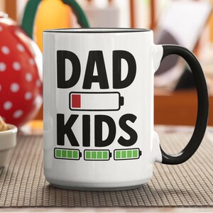 Peut inclure: Mug en céramique blanche avec anse et bord noir. Le mug affiche les mots "DAD" et "KIDS" en gras noir, avec des indicateurs de charge de batterie. Une théière rouge à pois blancs et un bol de biscuits sont en arrière-plan.