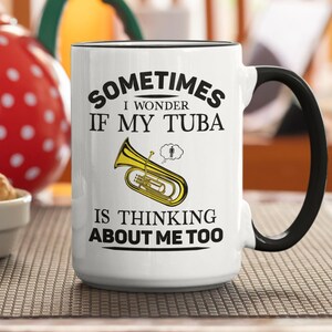 Peut inclure: Mug en céramique blanche avec anse et bord noir. Le mug porte l'inscription "Sometimes I wonder if my tuba is thinking about me too" et une illustration de tuba. Le mug est sur une surface marron.