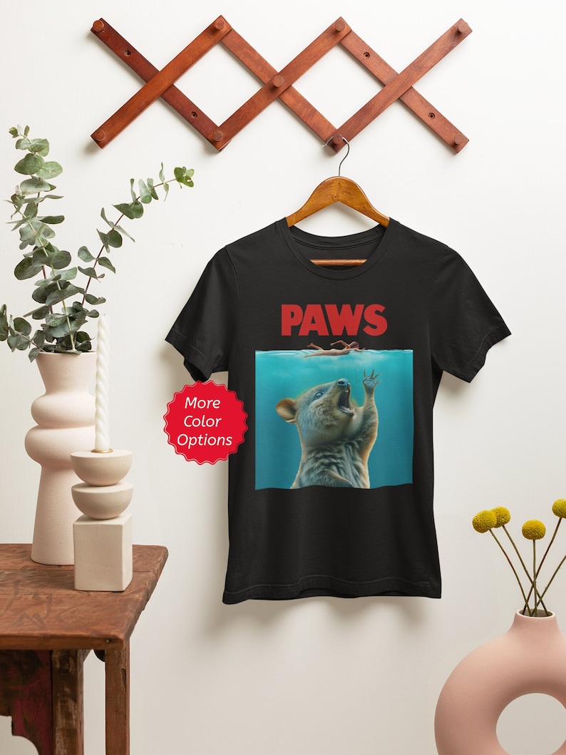 Puede incluir: Camiseta negra con un dise&ntilde;o parodia que presenta la palabra "PAWS" en rojo, sobre una ilustraci&oacute;n de un quokka en el agua, extendiendo la mano hacia un nadador. El dise&ntilde;o es una versi&oacute;n humor&iacute;stica de un cartel de pel&iacute;cula cl&aacute;sico.