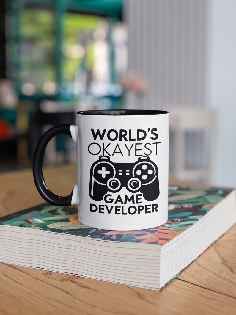 May include: White coffee mug with a black rim and handle. The mug has a black and white graphic of a video game controller and the text "WORLD'S OKAYEST GAME DEVELOPER".