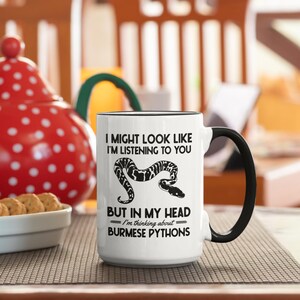 Op de afbeelding: Witte keramische mok met een zwart handvat en rand, met de tekst "I MIGHT LOOK LIKE I'M LISTENING TO YOU BUT IN MY HEAD I'm thinking about BURMESE PYTHONS" en een slangillustratie. De mok staat op een patroon oppervlak.